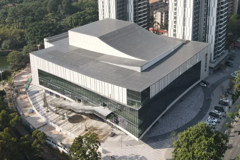 Aerial view of the completed Maoming City Theatre in China, showing the curved roof line and the modern glass-and-stone facade surrounded by parkland and high-rise context