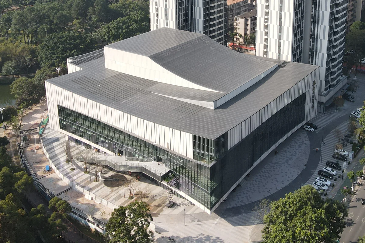 Aerial view of the completed Maoming City Theatre in China, showing the curved roof line and the modern glass-and-stone facade surrounded by parkland and high-rise context