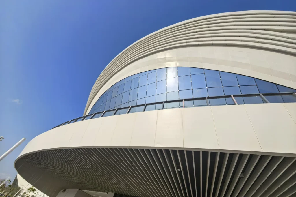 Completed curved sports arena in Botswana with glass curtain wall and three-dimensional aluminium panel facade against a clear blue sky