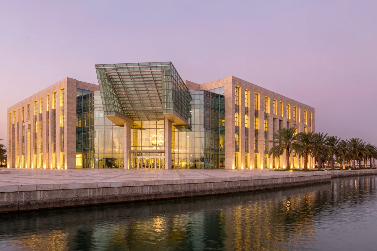 Four-storey performing arts centre on the Basrah waterfront with a unitized curtain wall glazed lobby glowing at dusk and a cantilevered entrance canopy over the plaza